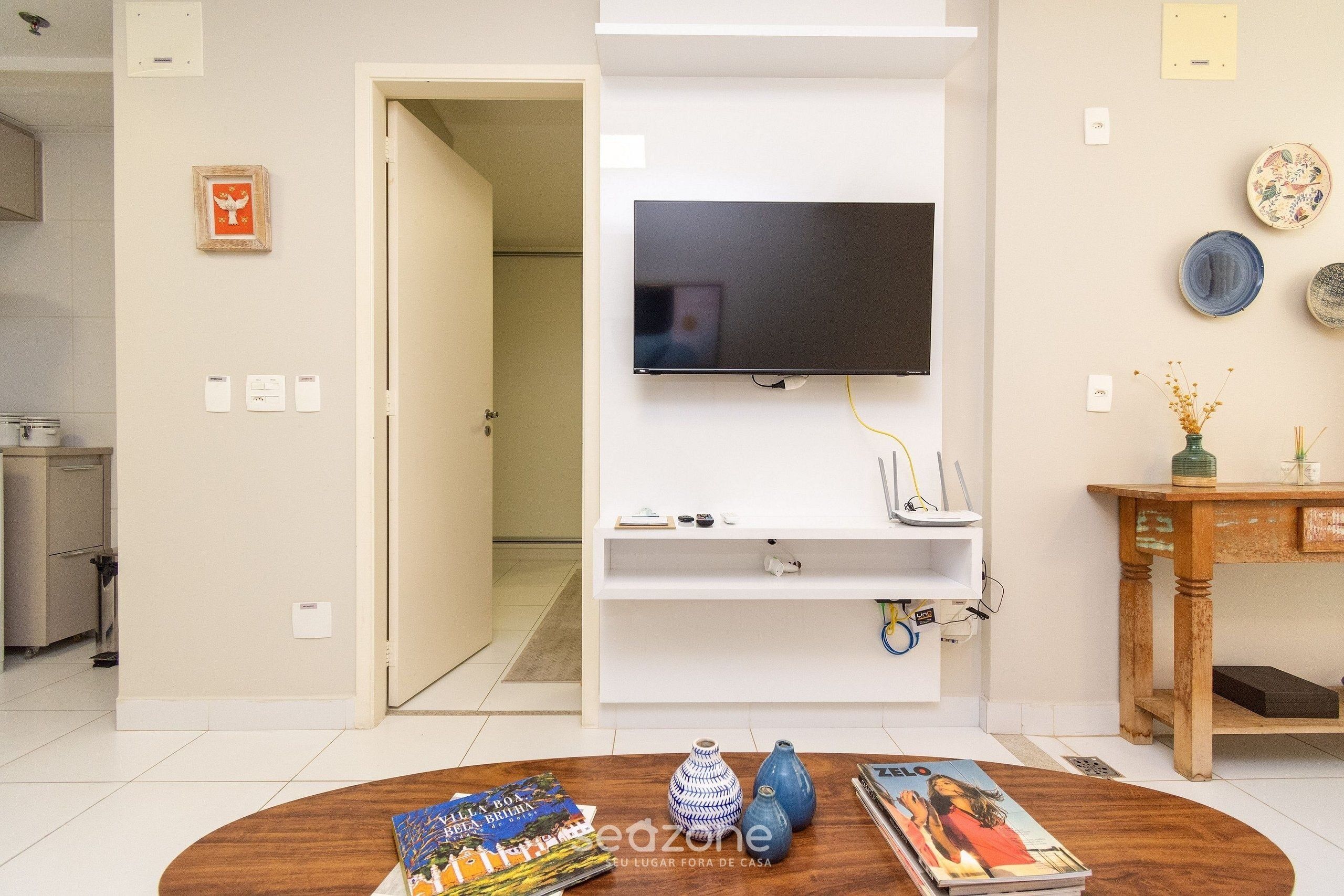 Una sala de estar con un televisor de pantalla plana montado en una pared blanca, una mesa de centro de madera con libros y jarrones, y una pequeña mesita auxiliar con objetos decorativos.