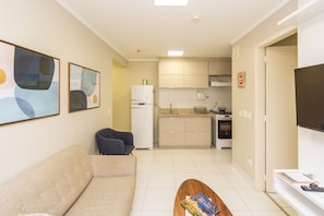 Una sala de estar con sofá, mesa de centro y televisor. Cuenta con una zona de cocina con armarios y un refrigerador.