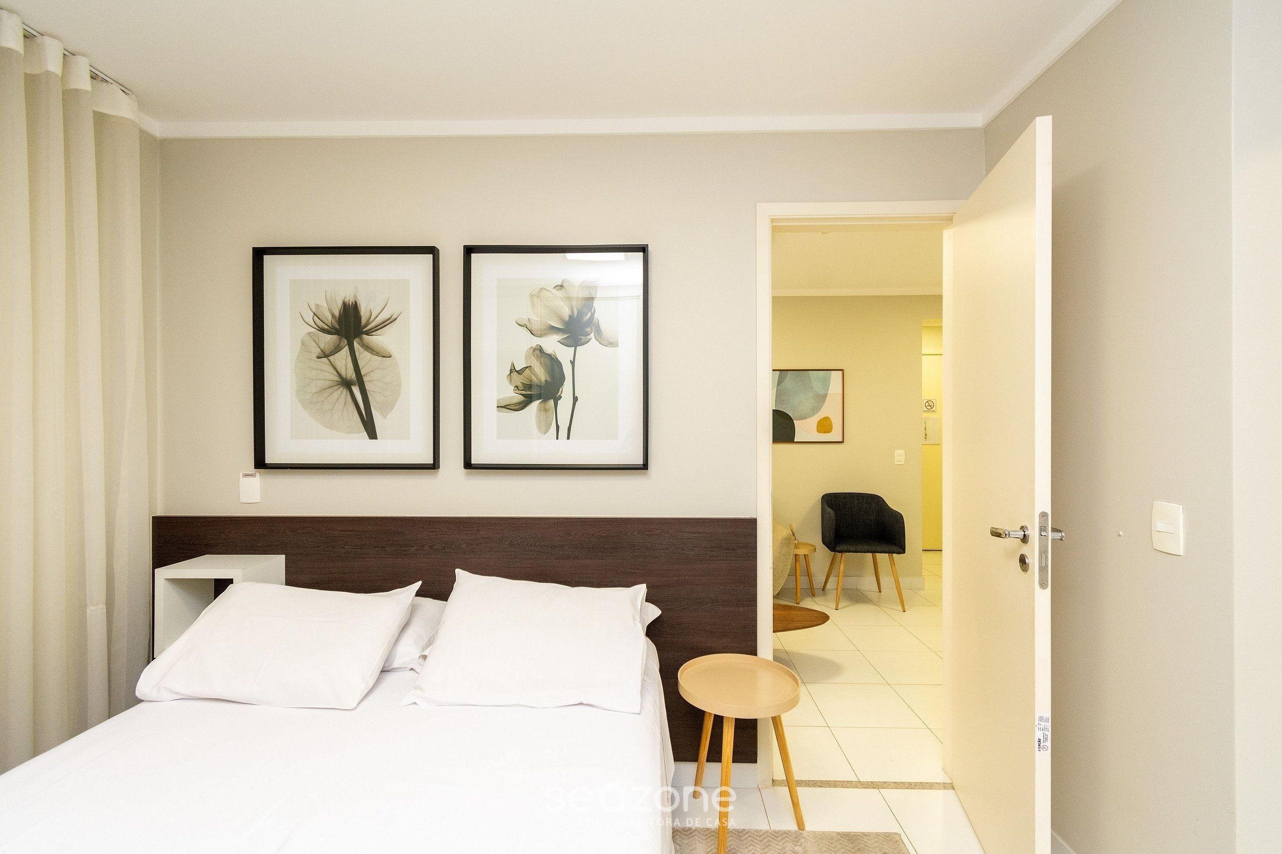 Habitación de hotel con una cama, dos cuadros con flores en la pared, una silla y una puerta que da a otra habitación.