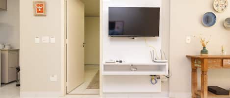 Een woonkamer met een flatscreen televisie aan de muur, een houten salontafel met tijdschriften en vazen, en een keuken in de achtergrond.