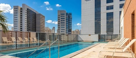 A rooftop pool with lounge chairs, a glass railing, and a cityscape view.