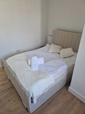 A neatly made bed with white linens and pillows, a wooden floor, and a plain white wall.