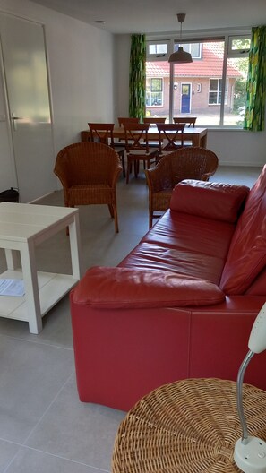 A red leather sofa, wicker chairs, a white table, and a dining area with a view of a red-roofed building. - Panda,6-person cottage at the foot of the dunes, a 5-minute walk from the beach. (Vrouwenpolder)