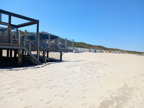 A beach with a wooden structure with steps leading to a covered area where people are seated. - Panda,6-person cottage at the foot of the dunes, a 5-minute walk from the beach. (Vrouwenpolder)