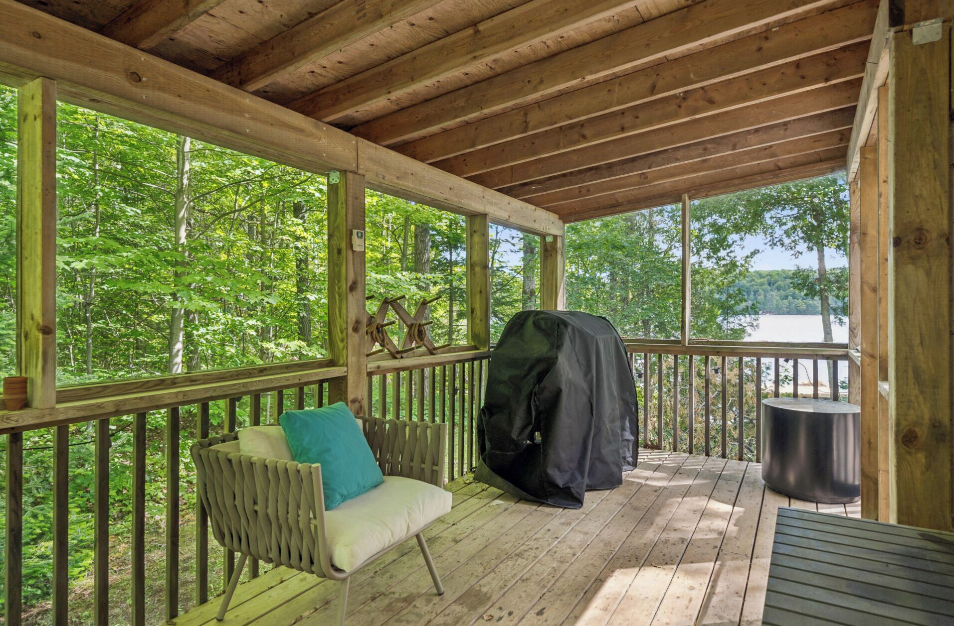 A covered outdoor area with a wooden deck, a bench, a grill, and a view of a lake surrounded by trees.