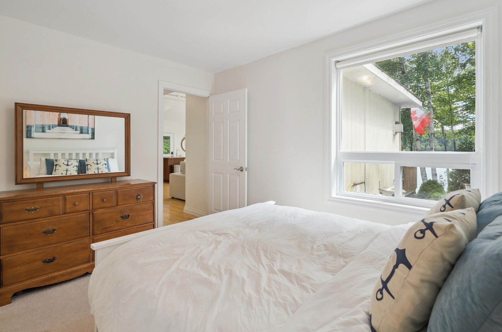 A bedroom with a bed, a dresser, a mirror, and a window with a view of trees and a flag.