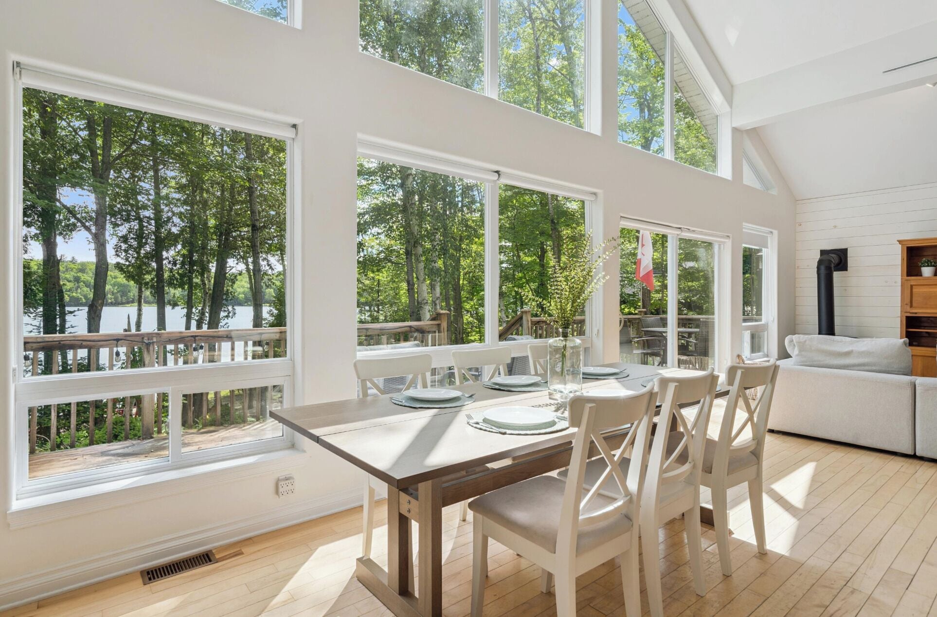 Un coin repas avec une table dressée pour quatre personnes, de grandes fenêtres offrant une vue sur les arbres et un plan d’eau, et un canapé blanc placé dans un coin.