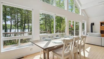 Un coin repas avec une table dressée pour quatre personnes, de grandes fenêtres offrant une vue sur les arbres et un plan d’eau, et un canapé blanc placé dans un coin.