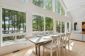 A dining area with a table set for four, large windows with a view of trees and a body of water, and a white sofa in the corner.