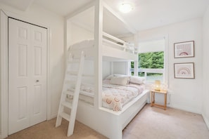 A bunk bed room with a ladder, a bedside table, and a window with a view of greenery.