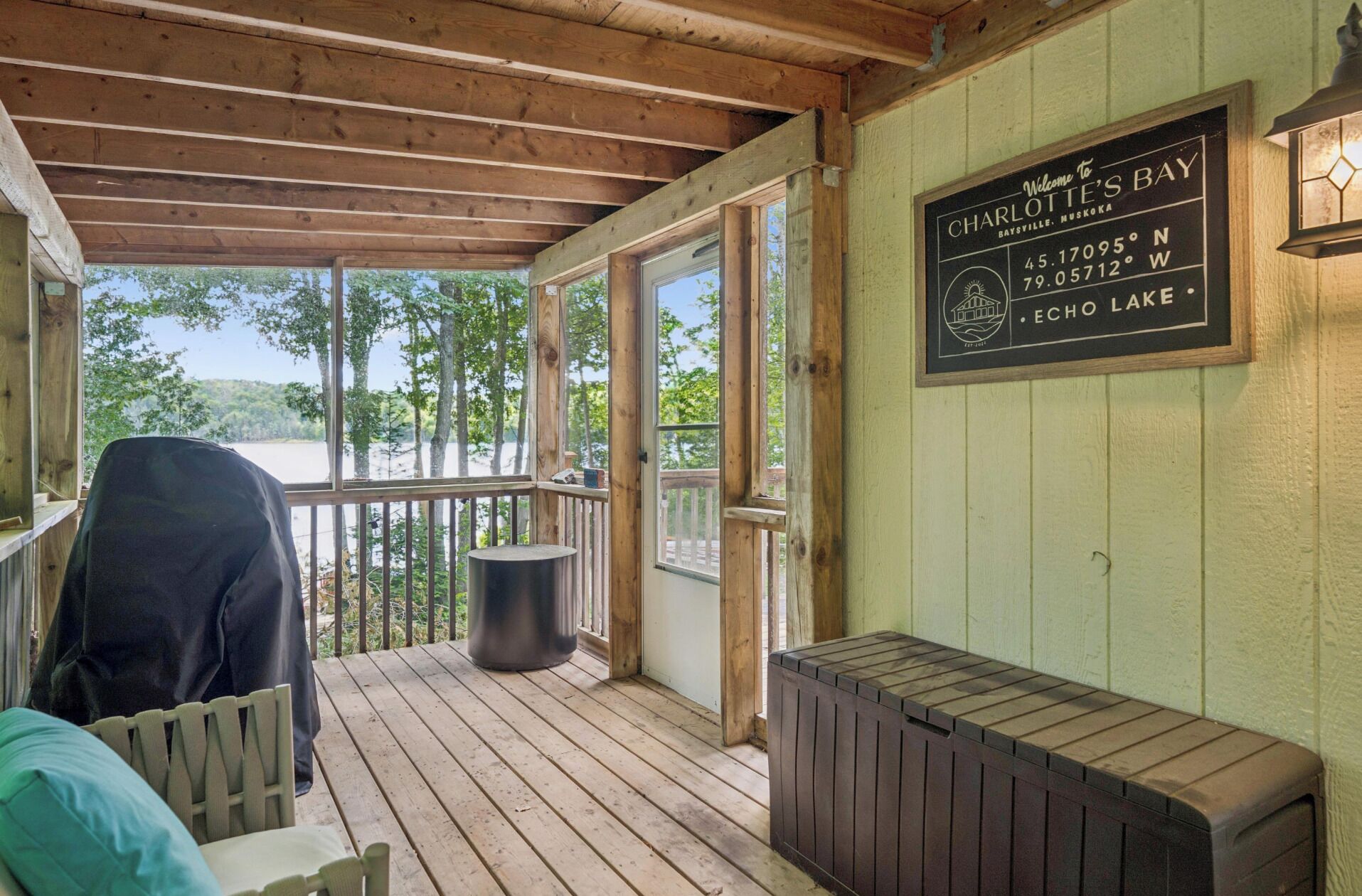 Une terrasse en bois dotée d’un espace salon couvert, une pancarte indiquant qu’on se trouve à Charlotte’s Bay, et une vue sur les arbres et l’eau environnants.