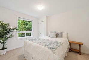 Un dormitorio con cama, una banca, una planta en maceta y una ventana con vista a follaje.