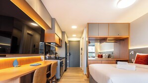 A modern hotel room with a kitchenette, a bed with orange pillows, and a view of the city. - Ária Higienópolis by WeCare Hosting (São Paulo)