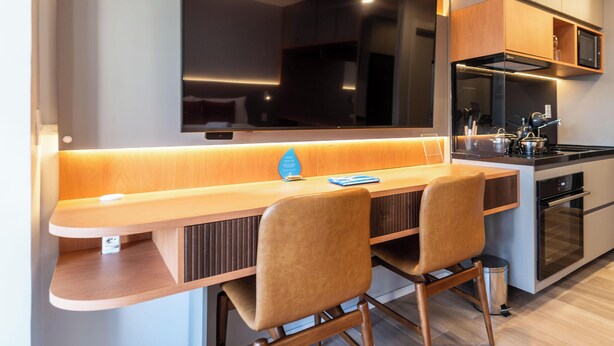 A modern kitchen with a built-in TV, wooden bar counter, two chairs, and stainless steel appliances. - Ária Higienópolis by WeCare Hosting (São Paulo)