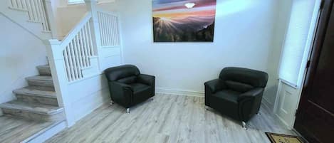A room with a wooden floor, two black armchairs, a staircase with white railings, and a painting on the wall.