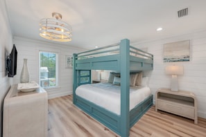 Uma cama beliche com colchão branco e estrutura azul-esverdeada, uma mesinha de cabeceira com abajur e uma TV fixada na parede.