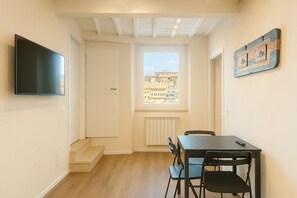 A narrow room with a TV mounted on the wall, a window with a view of buildings, a table with chairs, and a radiator. - Top Floor - Top Location (Siena)