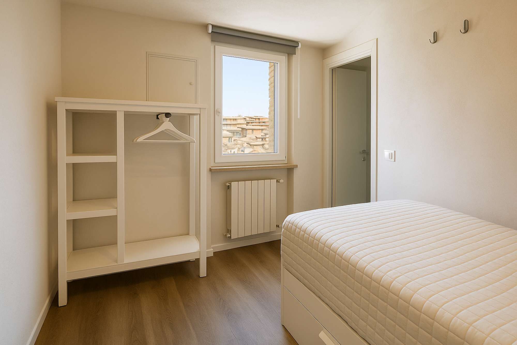 A bedroom with a bed, a wardrobe, a radiator, and a window with a view of buildings.