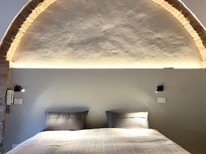 A bedroom with a brick arch ceiling, a bed with striped bedding, two bedside lamps, and a wall-mounted light switch. - Very Central Studio close to public parking and il Campo (Siena)