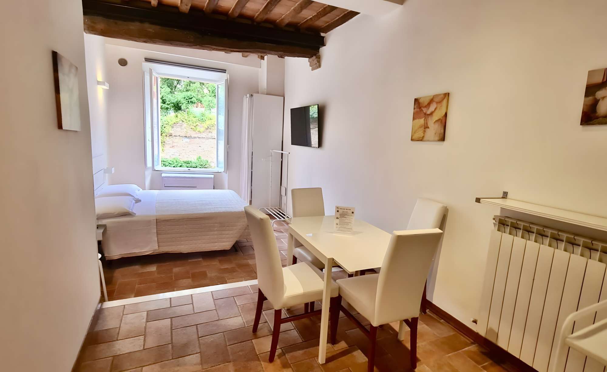 A compact room with a bed, a table with chairs, a radiator, and a window with a view of greenery.