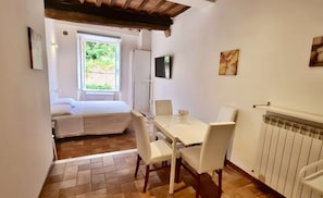 A compact room with a bed, a table with chairs, a radiator, and a window with a view of greenery. - Very central studio II close to public parking and il Campo (Siena)