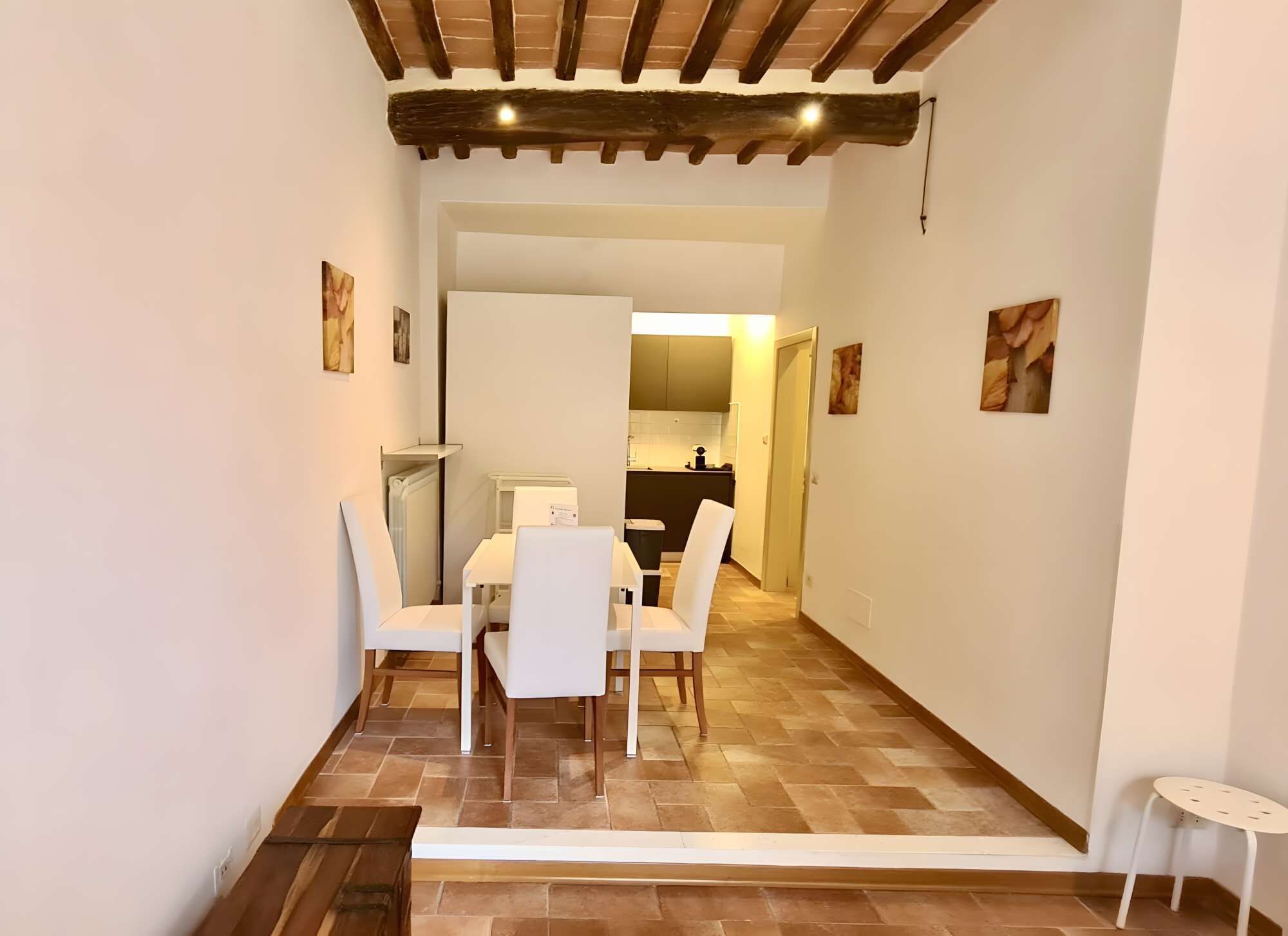 A dining area with a table and chairs, a kitchen in the background, and a wooden ceiling.