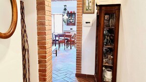 A hallway with a brick column, a dining area with a table and chairs, and a wooden cabinet with glassware. - A BALCONY ON THE PIAZZA OF THE PALIO (Siena)