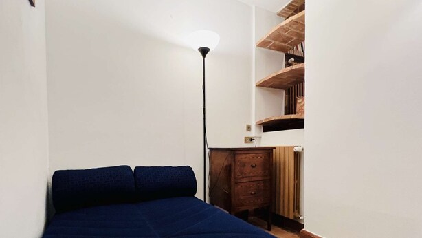 A bedroom with a bed, a floor lamp, a wooden dresser, and a brick-patterned shelf. - A BALCONY ON THE PIAZZA OF THE PALIO (Siena)