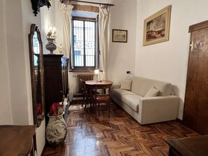 Ein Zimmer mit Holzdielen-Fußboden, einem beigen Sofa, einem kleinen runden Tisch, einem Spiegel mit dekorativem Rahmen und einem Fenster mit Gitterstäben.