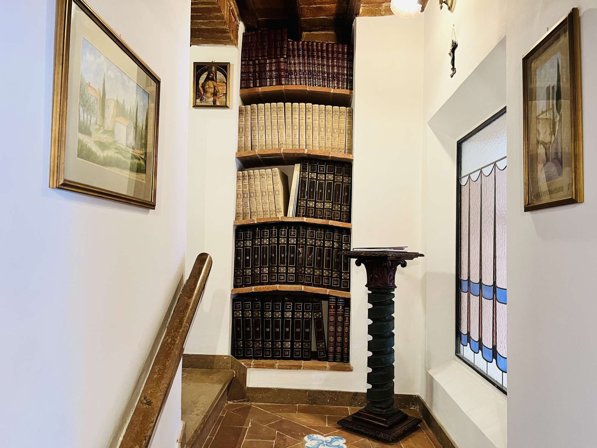Un couloir étroit, avec une bibliothèque remplie de livres, un tableau encadré sur le côté gauche et un vitrail sur le côté droit.