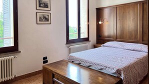 A bedroom with a bed, a wooden wardrobe, two framed pictures on the wall, and a window with a view of greenery. - Cozy Studio 8 in the Heart of Siena (Siena)