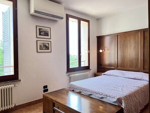 Ein Schlafzimmer mit einem Bett, einem Holzschrank, zwei gerahmten Bildern an der Wand und einem Fenster mit Blick auf Grünflächen.