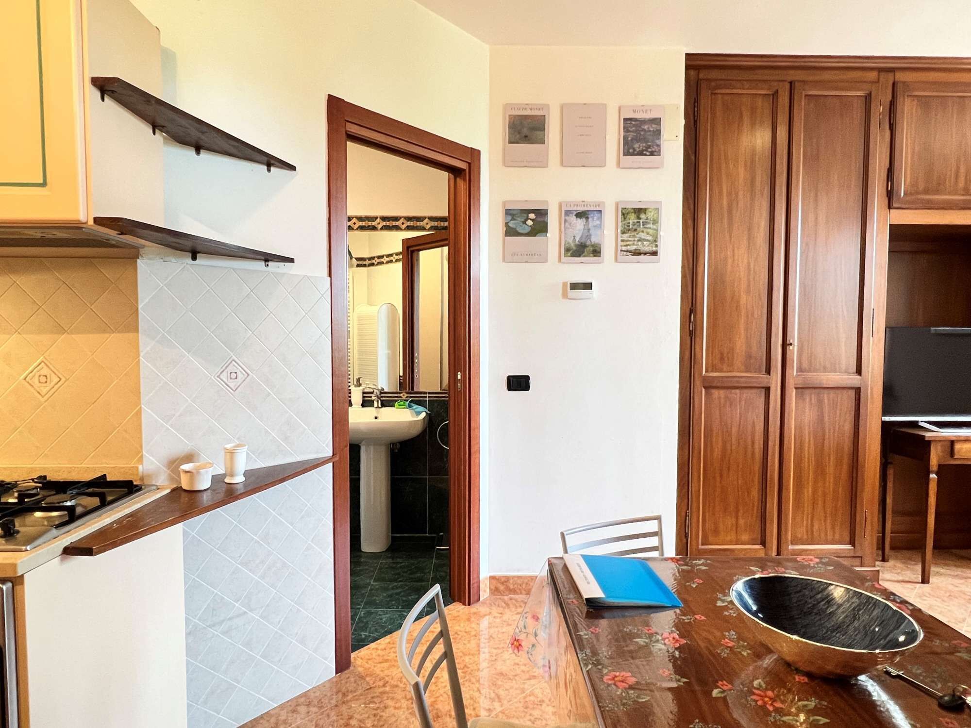 Una cucina con piano cottura a gas, lavello e pensili in legno. Si nota un tavolo da pranzo con sedie e una ciotola decorativa.