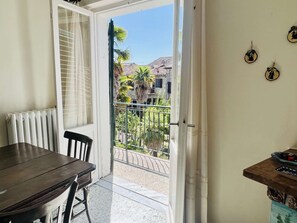 Una habitació amb taula i cadires, un radiador, una porta corredissa de vidre que dona a un balcó amb vistes a edificis i palmeres.