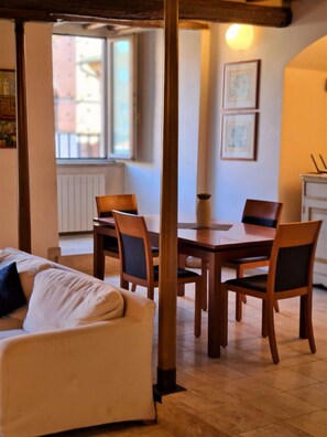 A dining area with a wooden table and chairs, a sofa, and a window with a view of buildings. - Apartment in Piazza del Campo area 3 bedrooms 2 bathrooms (siena)