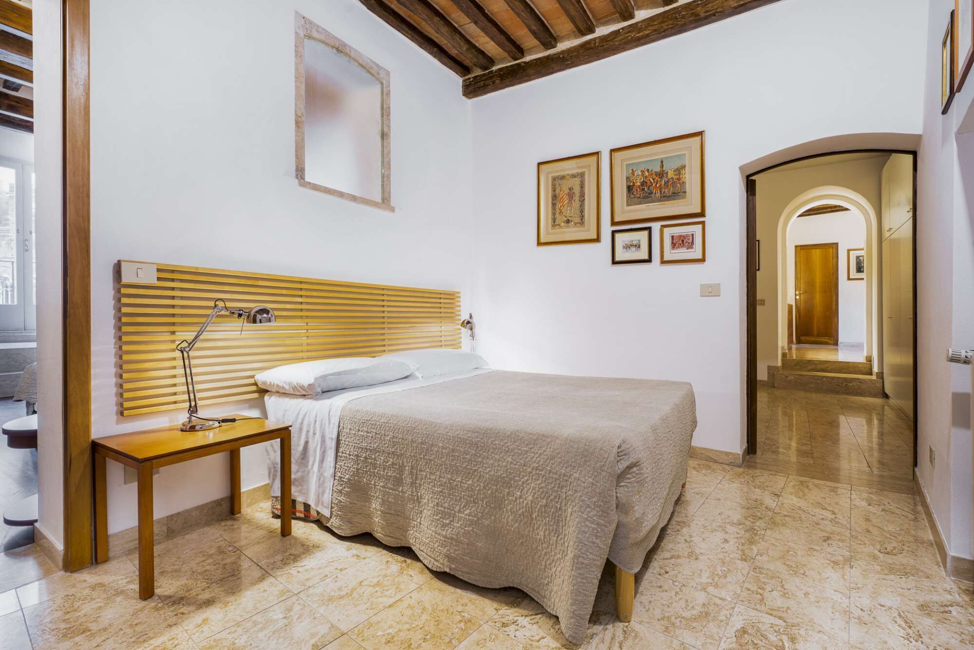 A bedroom with a wooden headboard, a bedside table with a lamp, a bed with a textured blanket, and a hallway leading to a door.