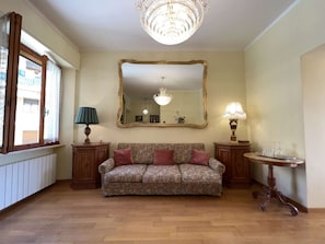 Ein Zimmer mit einer Couch, zwei Beistelltischen, einem Spiegel, einer Chandelüster und einem fenster mit Vorhängen.