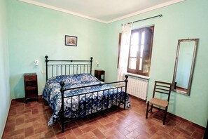 Una habitación con cama, una silla, ventana con cortinas, radiador y un espejo en la pared.