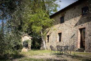 Un edificio de piedra rústico con una mesa y sillas en primer plano, un árbol a la izquierda y un cielo despejado.