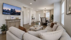 Una sala de estar moderna con un televisor de pantalla plana grande, un conjunto de sofás, una mesa de centro y un área de comedor al fondo.