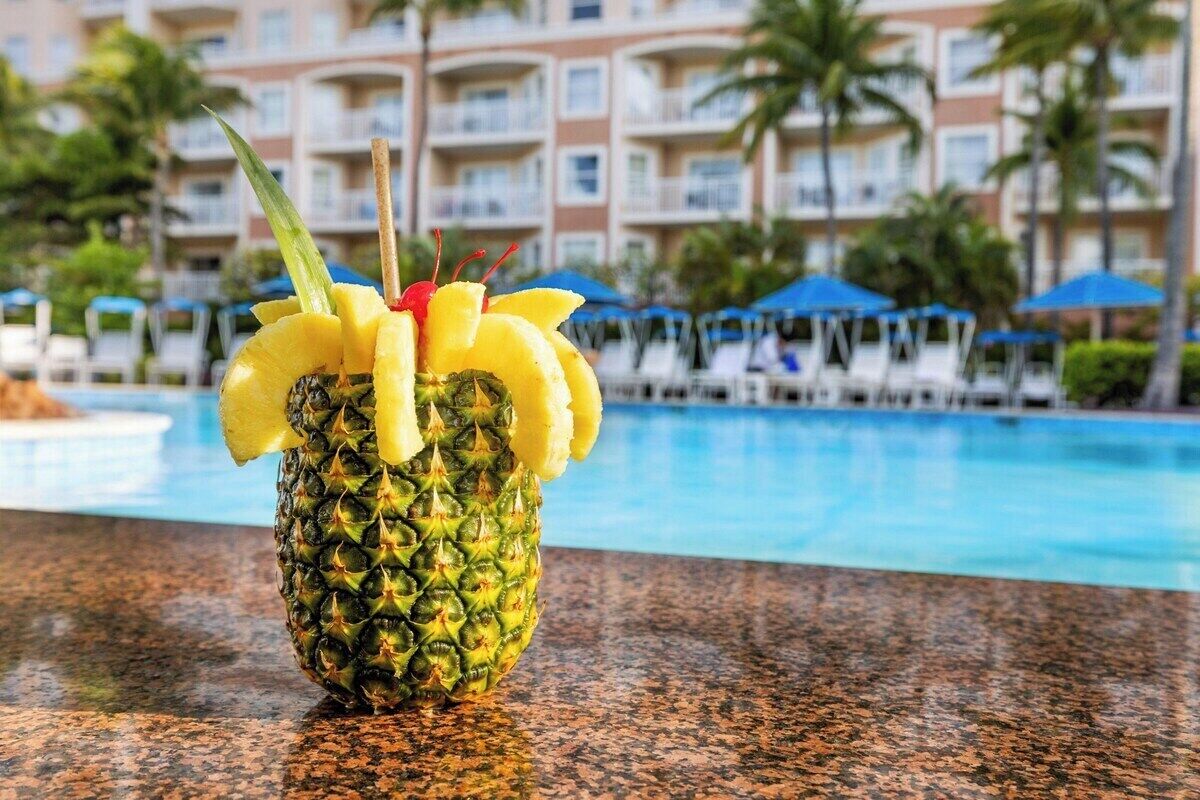 Cóctel de piña adornado con cereza, sobre una encimera de granito cerca de una piscina, con un edificio de hotel al fondo.