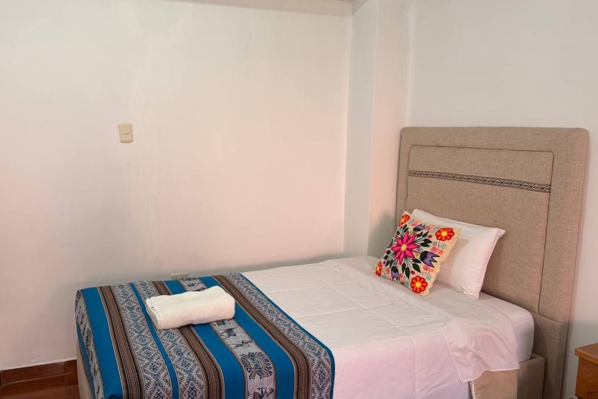 A single bed with a patterned bedspread and a decorative pillow, a headboard, and a small bedside table with a lamp.