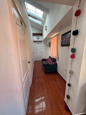 Un couloir étroit avec un plancher en bois, une porte blanche et un canapé bleu agrémenté d’un plaid à motifs.
