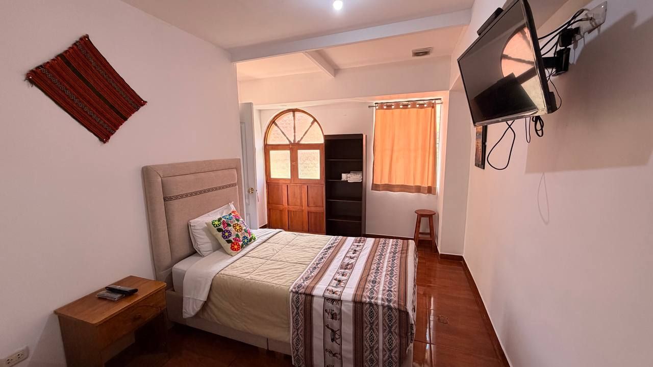Un dormitorio con cama, mesita de noche, televisor, armario y ventana con cortinas.