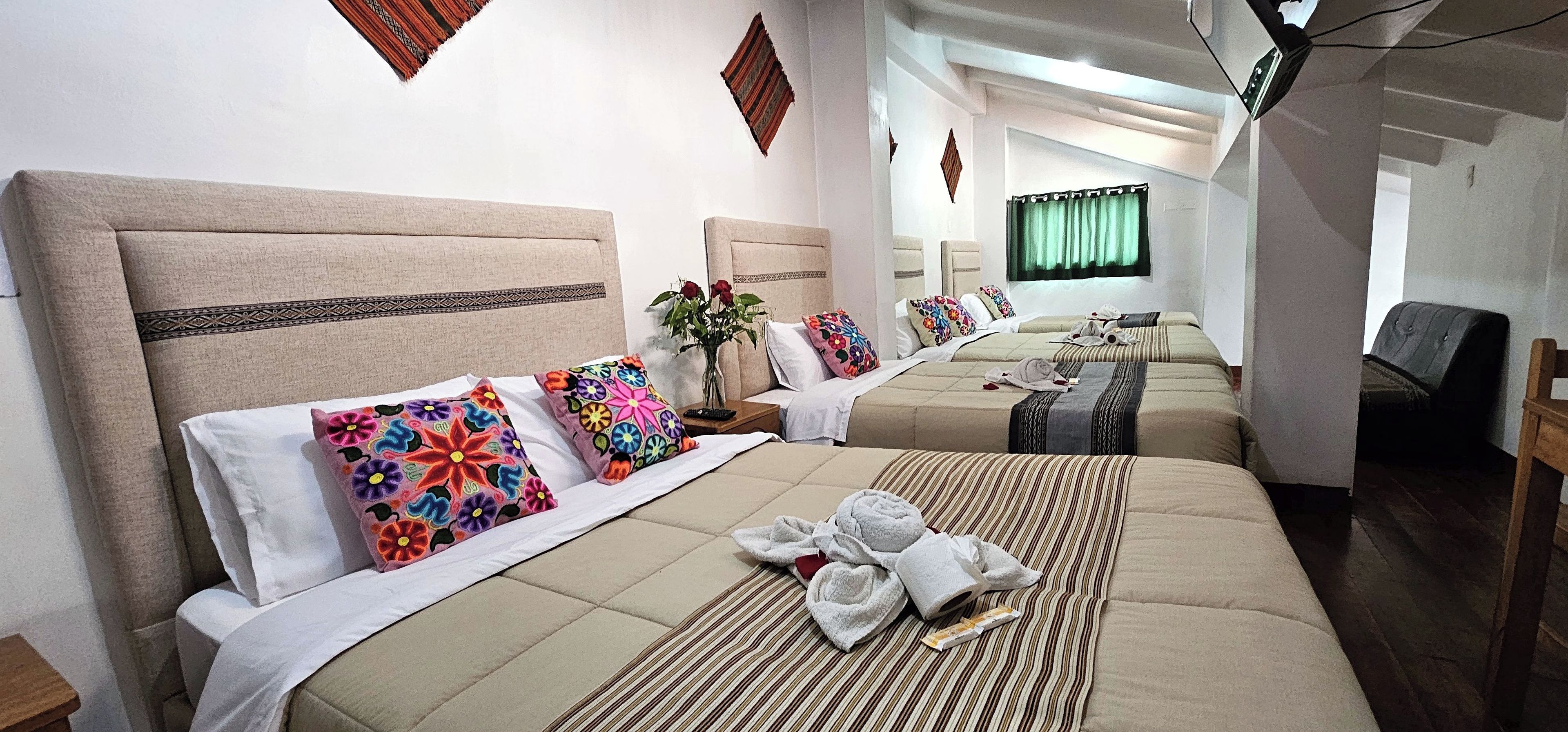 Quarto de hotel com três camas, cada uma com cabeceira e várias almofadas. H á uma mesinha com um vaso de flores e um tabuleiro com toalhas e um livro.
