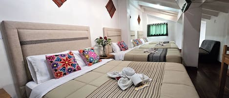 Habitación de hotel con tres camas, cada una con cabecera y varias almohadas. Una mesita pequeña con un florero y una bandeja con toallas y un libro.