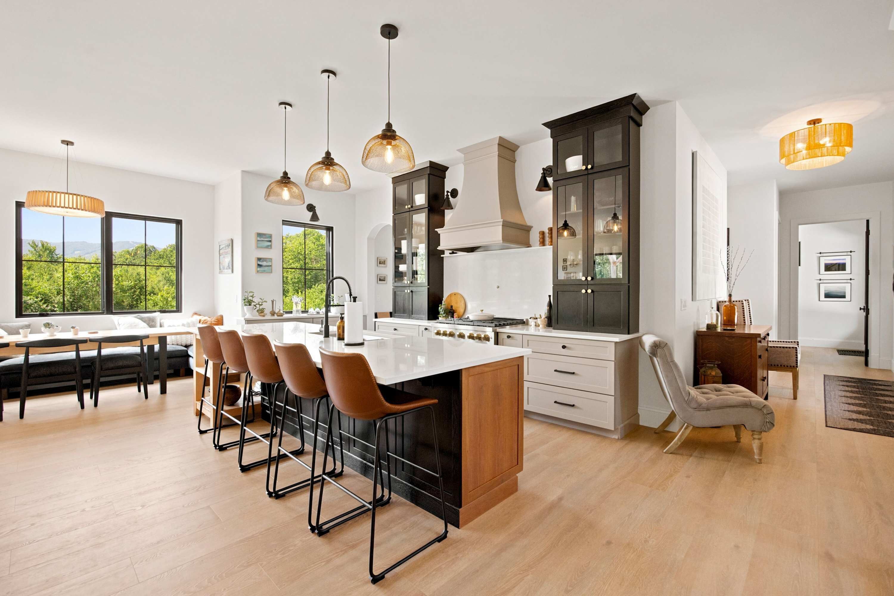 A modern kitchen with a central island, bar stools, pendant lighting, and a view of greenery outside.