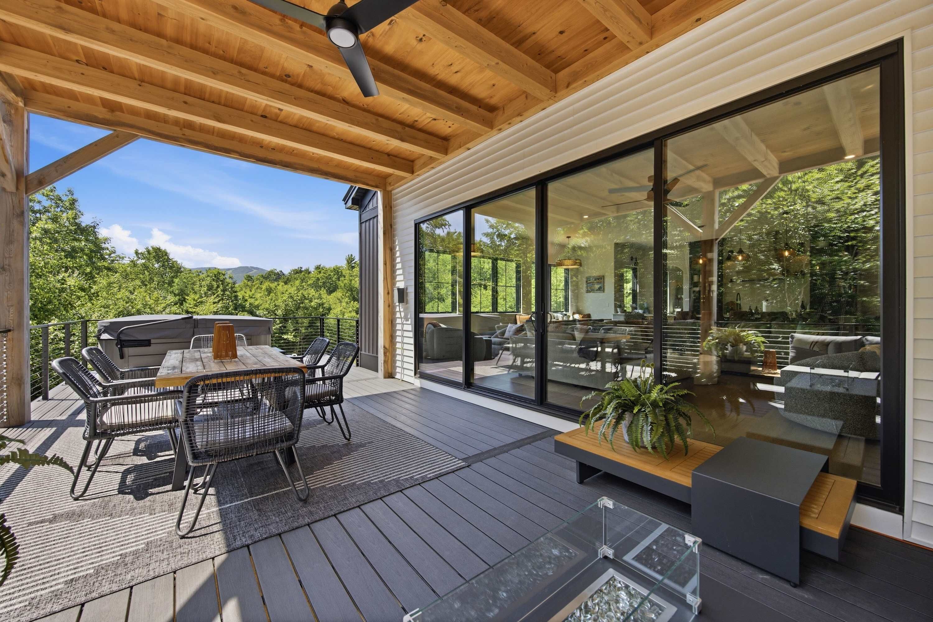 Una terraza exterior con techo de madera, una mesa de comedor con sillas, una parrilla y un amplio ventanal corredero de vidrio que da a una zona de estar interior.