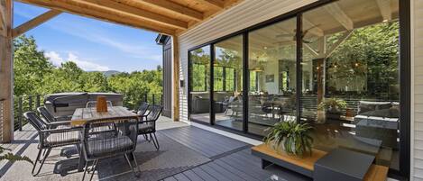 Una terraza exterior con techo de madera, una mesa de comedor con sillas, una parrilla y un amplio ventanal corredero de vidrio que da a una zona de estar interior.