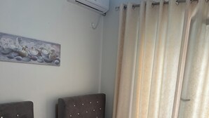 A hotel room with two beds, a wall-mounted air conditioner, a painting, and a window with curtains. - Cozy 1-bedroom apartment with sea view (Saranda)
